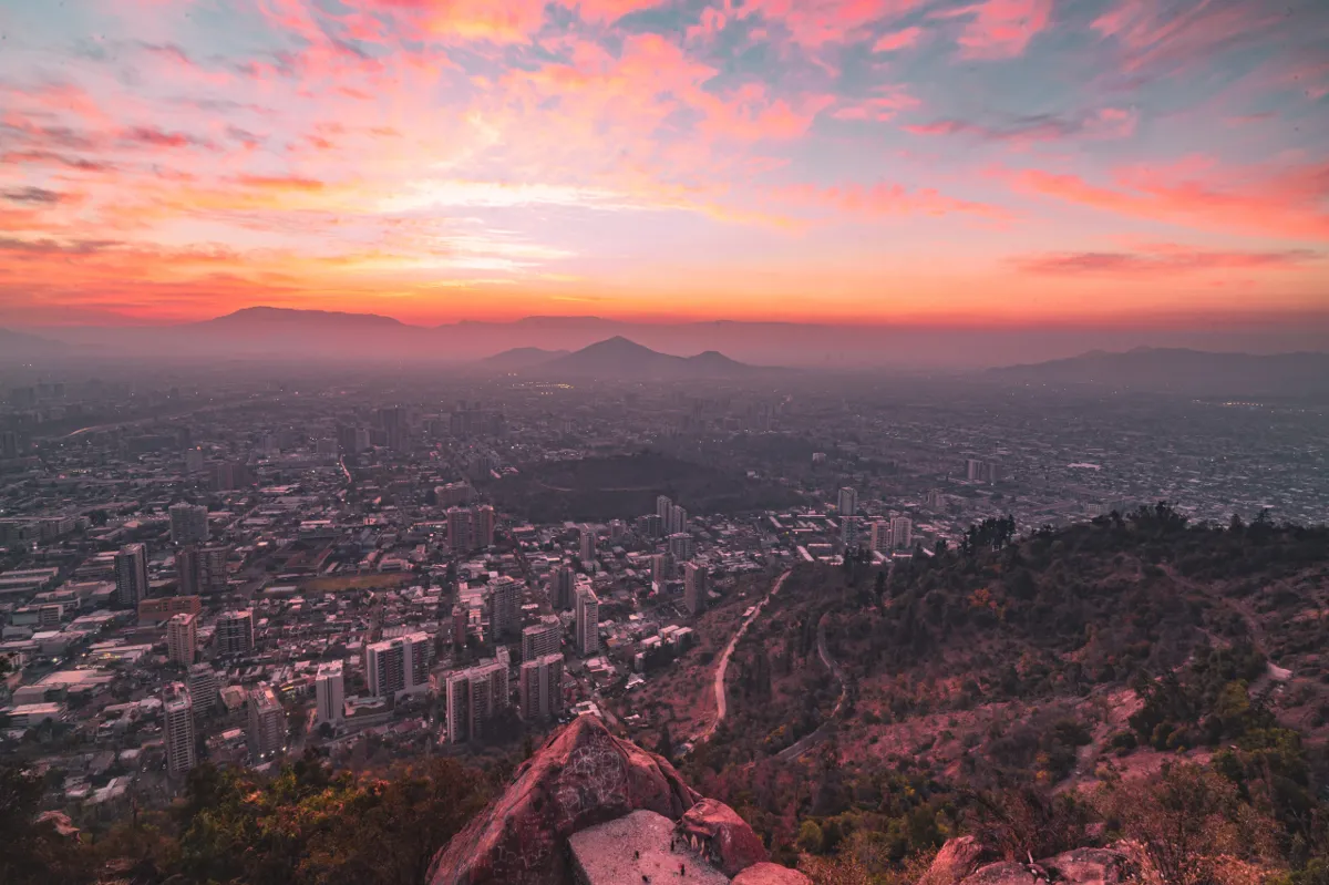 A vivid pink and orange sunset sky over the sprawling city of Santiago, seen from a rocky hilltop.