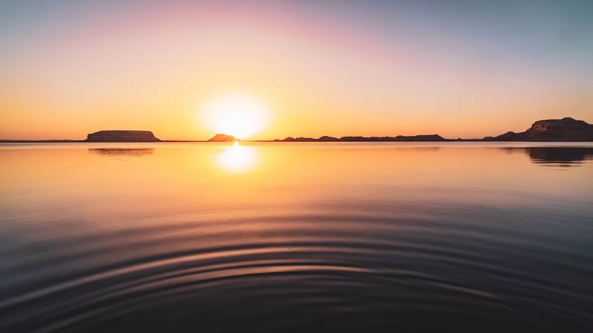 The sun sets over a still salt lake near Siwa, with ripples spreading across the water and flat-topped hills beyond.