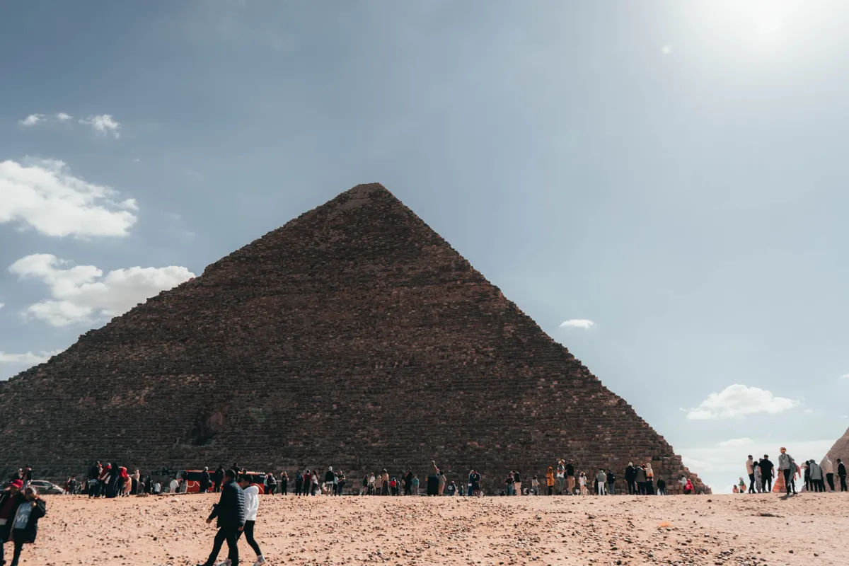 The Great Pyramid of Giza rises above a crowd of visitors on the sand, under a hazy sun.