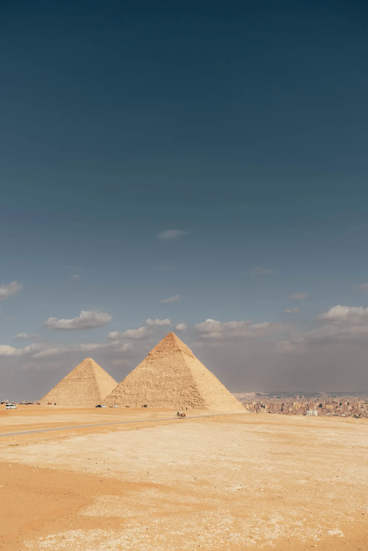 Two of the Giza pyramids stand on the desert plateau under a deep blue sky, with Cairo's haze beyond.