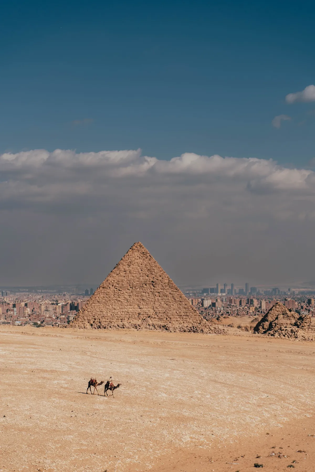 Two people on camels cross the desert below a Giza pyramid, with the Cairo skyline stretching behind.