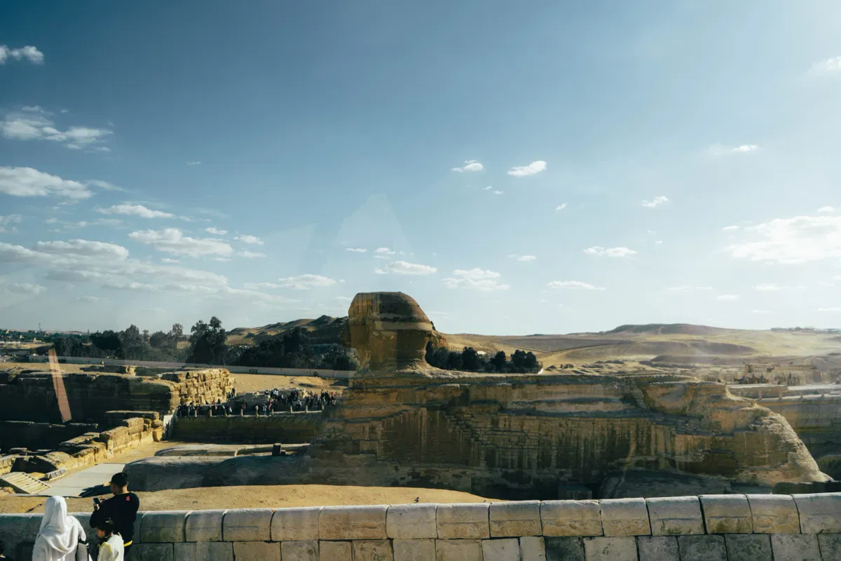 The Great Sphinx of Giza seen from a stone terrace, with visitors gathered below and desert hills beyond.