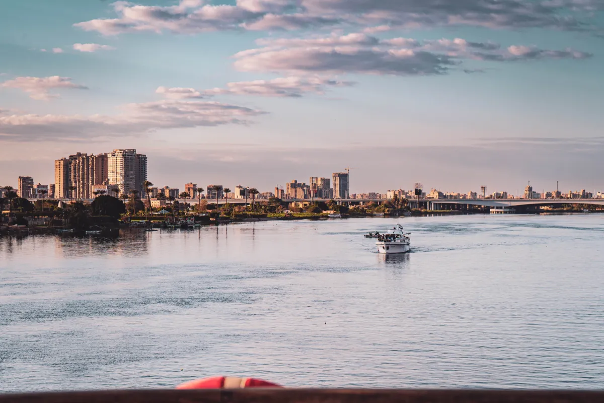 A small white boat crosses the Nile toward a bridge and the Cairo skyline under a pastel evening sky.