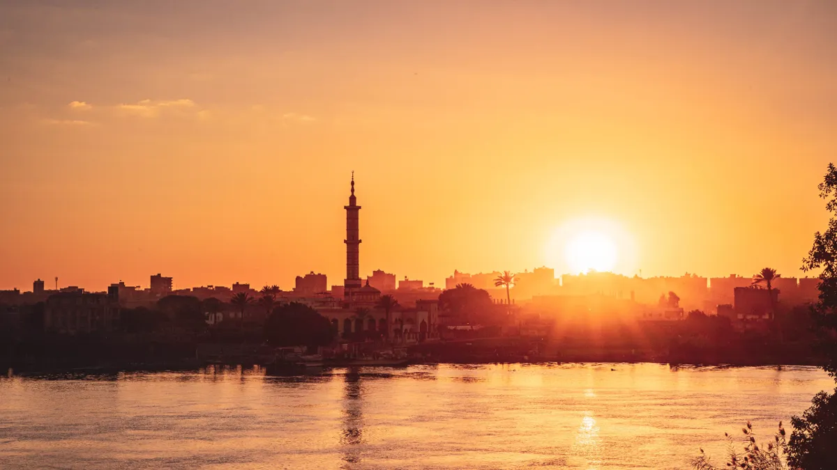 The sun sets in an orange sky behind a mosque minaret and palm trees, with the Nile in the foreground.