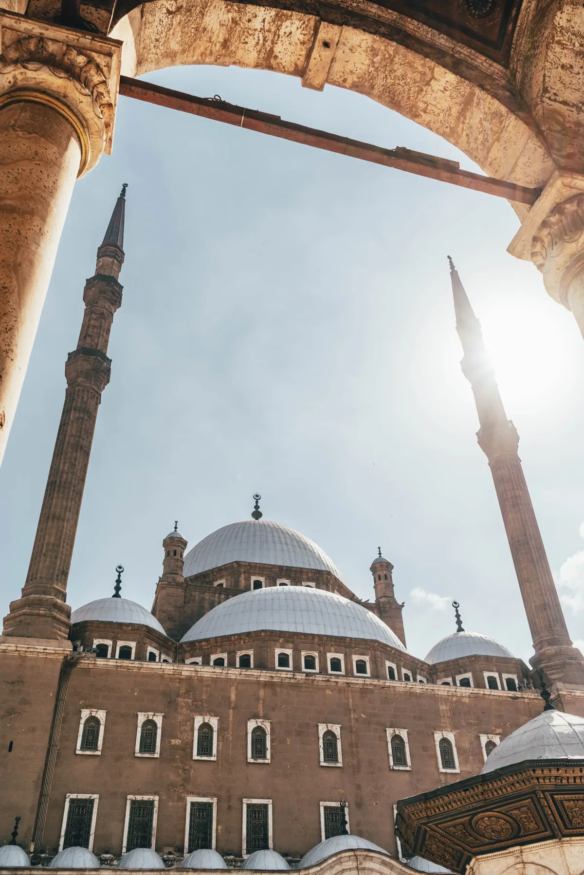 Two pencil-shaped minarets and domes of a Cairo mosque seen through a stone arch, with sunlight flaring at right.