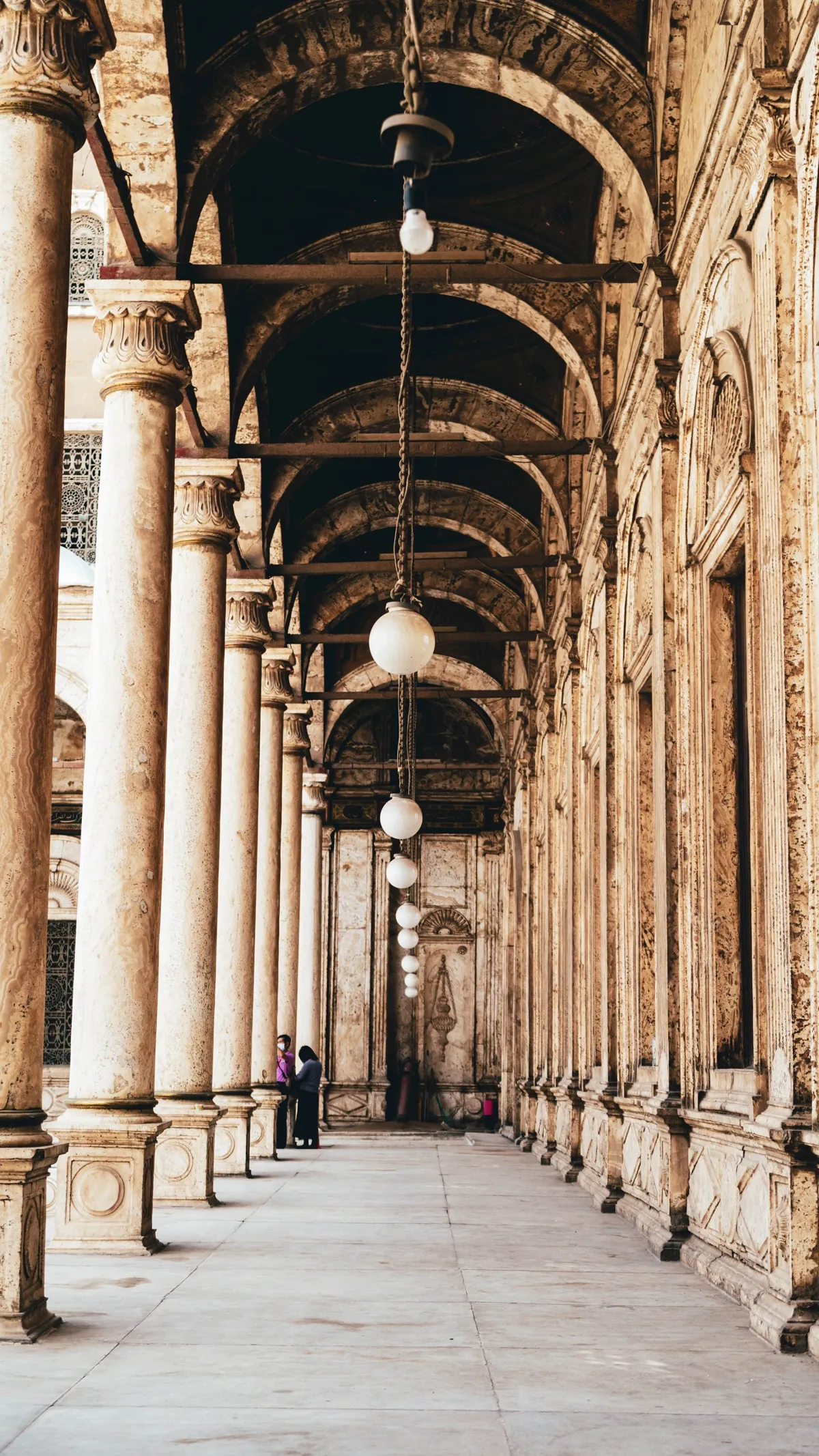 A stone colonnade in a mosque courtyard with hanging globe lamps and two people standing in the distance.