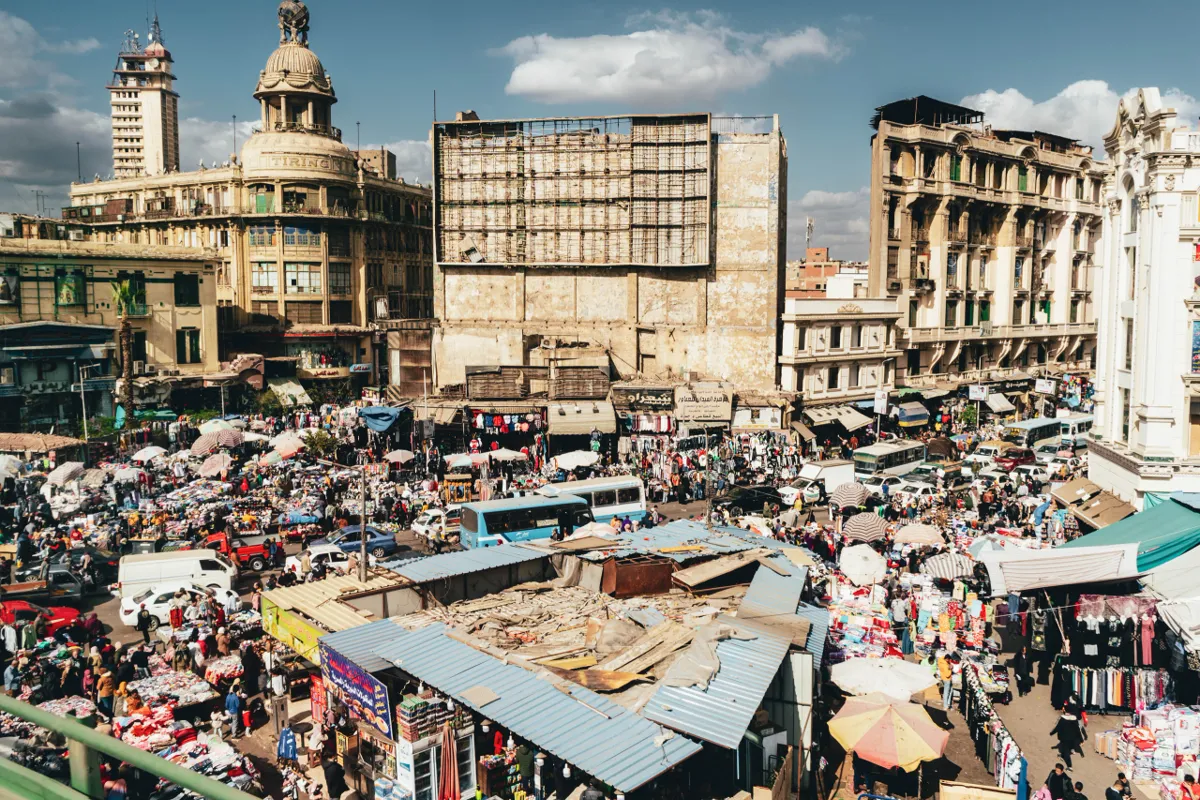 A crowded Cairo street market with awnings, buses and umbrellas below ornate old buildings under a blue sky.
