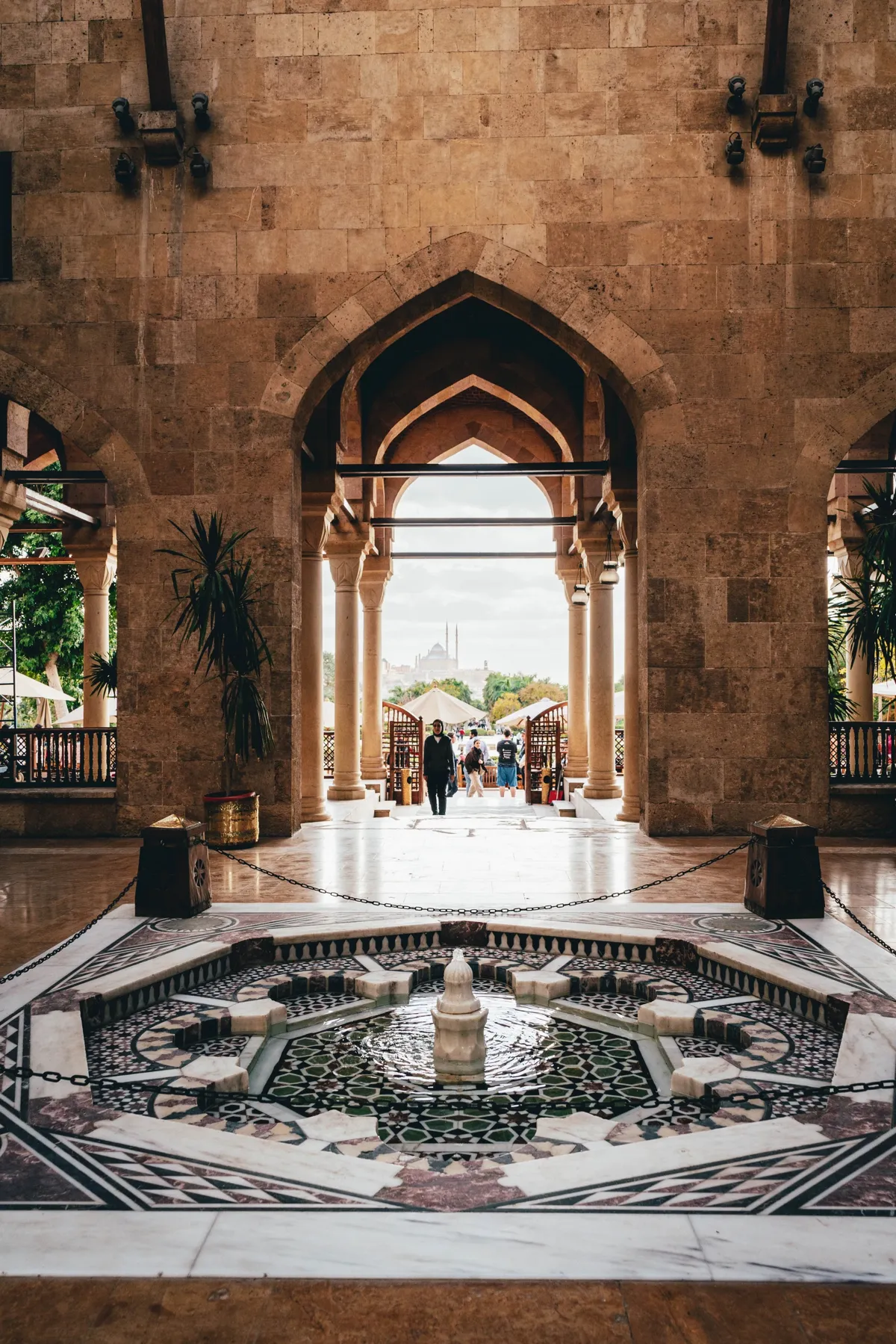 An ornate marble fountain inside a stone mosque hall, with arches opening onto a sunlit courtyard and a few visitors.