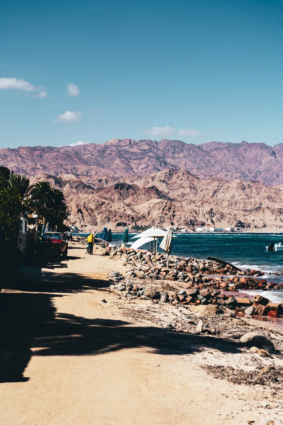 A sandy road beside a rocky Red Sea shore in Dahab, with rust-coloured mountains and a blue sky.
