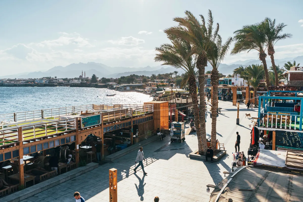 A seaside promenade in Dahab with wooden cafes, palm trees and people walking in the afternoon sun beside the bay.