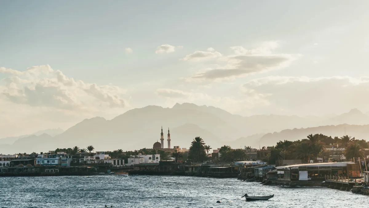 A mosque with two minarets on the Dahab shoreline in soft haze, with mountains behind and a small boat.