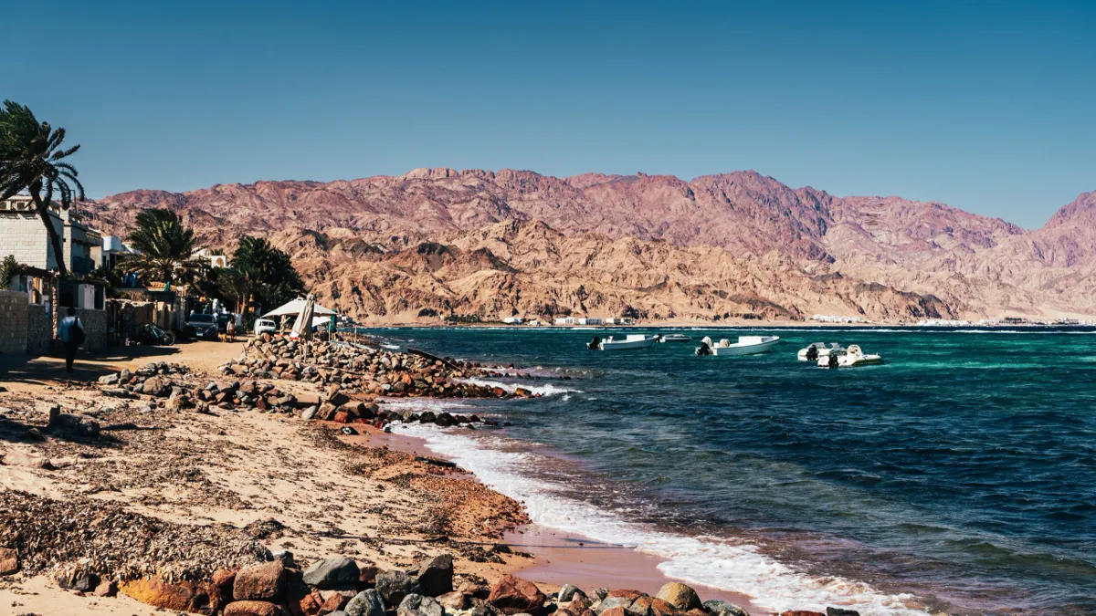 Small white boats on turquoise water along the rocky Dahab coastline, with red mountains rising behind.