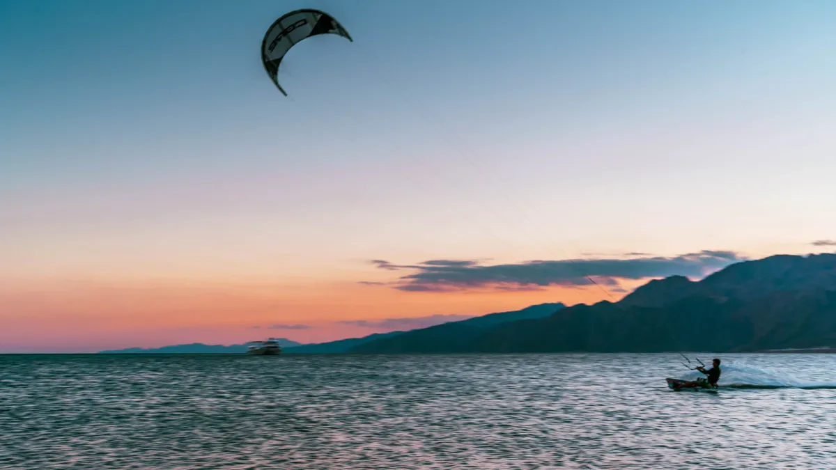 A kitesurfer skims across the Red Sea at sunset beneath a kite, with dark mountains and a pink sky.