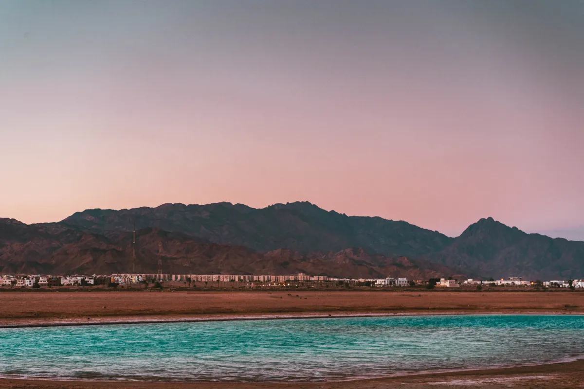 A turquoise lagoon in front of low buildings and rugged desert mountains under a pink dusk sky near Dahab.