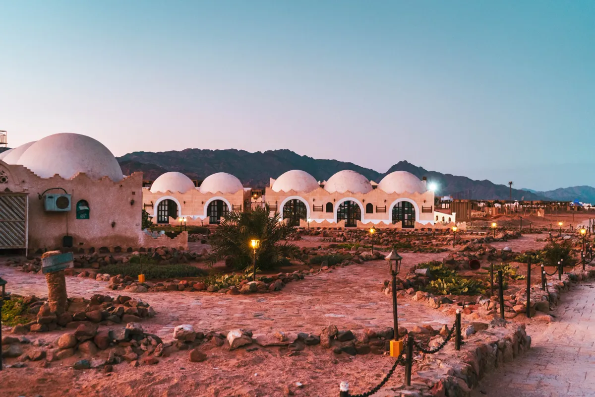 Domed adobe-style buildings and lanterns on a sandy path at dusk in Dahab, with mountains behind.