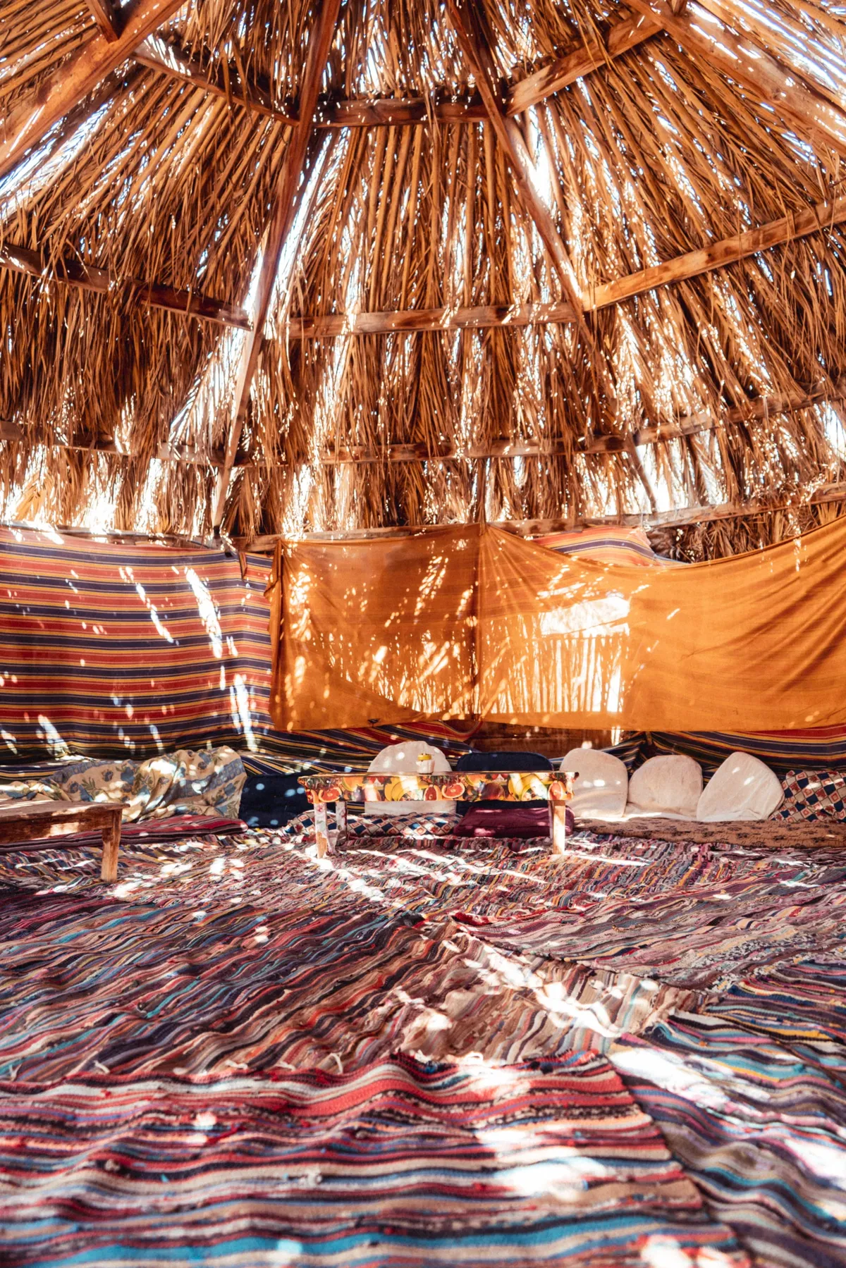 Inside a palm-thatched shelter in Dahab, sunlight falls through the roof onto striped woven rugs and cushions.