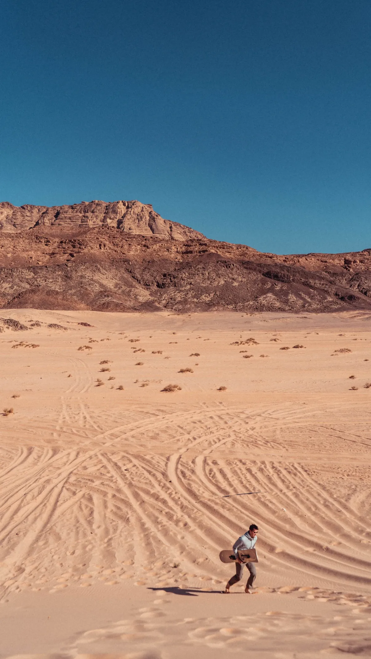 A person carrying a sandboard walks across rippled desert sand toward a rocky mountain under a deep blue sky.