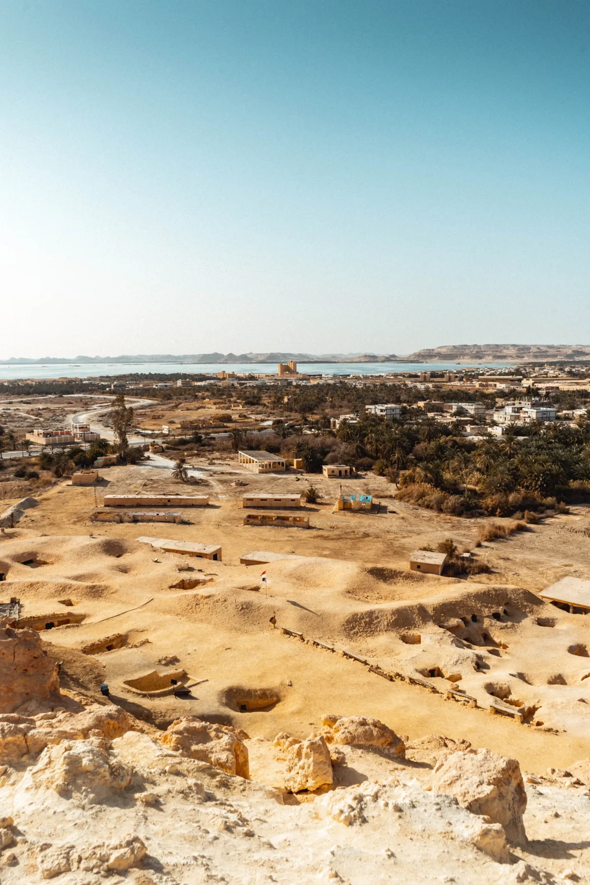 View over Siwa from a rocky hilltop, with sandy ruins in front and palm groves and a salt lake beyond.