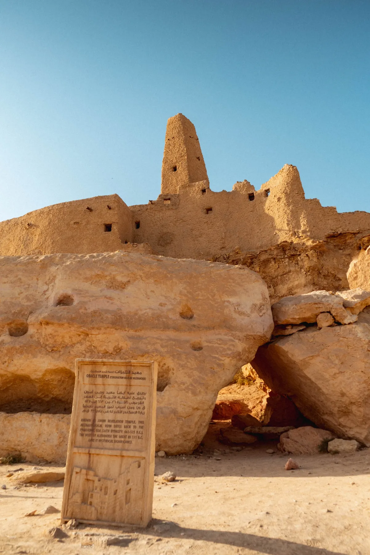 The mud-brick ruins of an ancient temple above large boulders in Siwa, with an information sign in front.