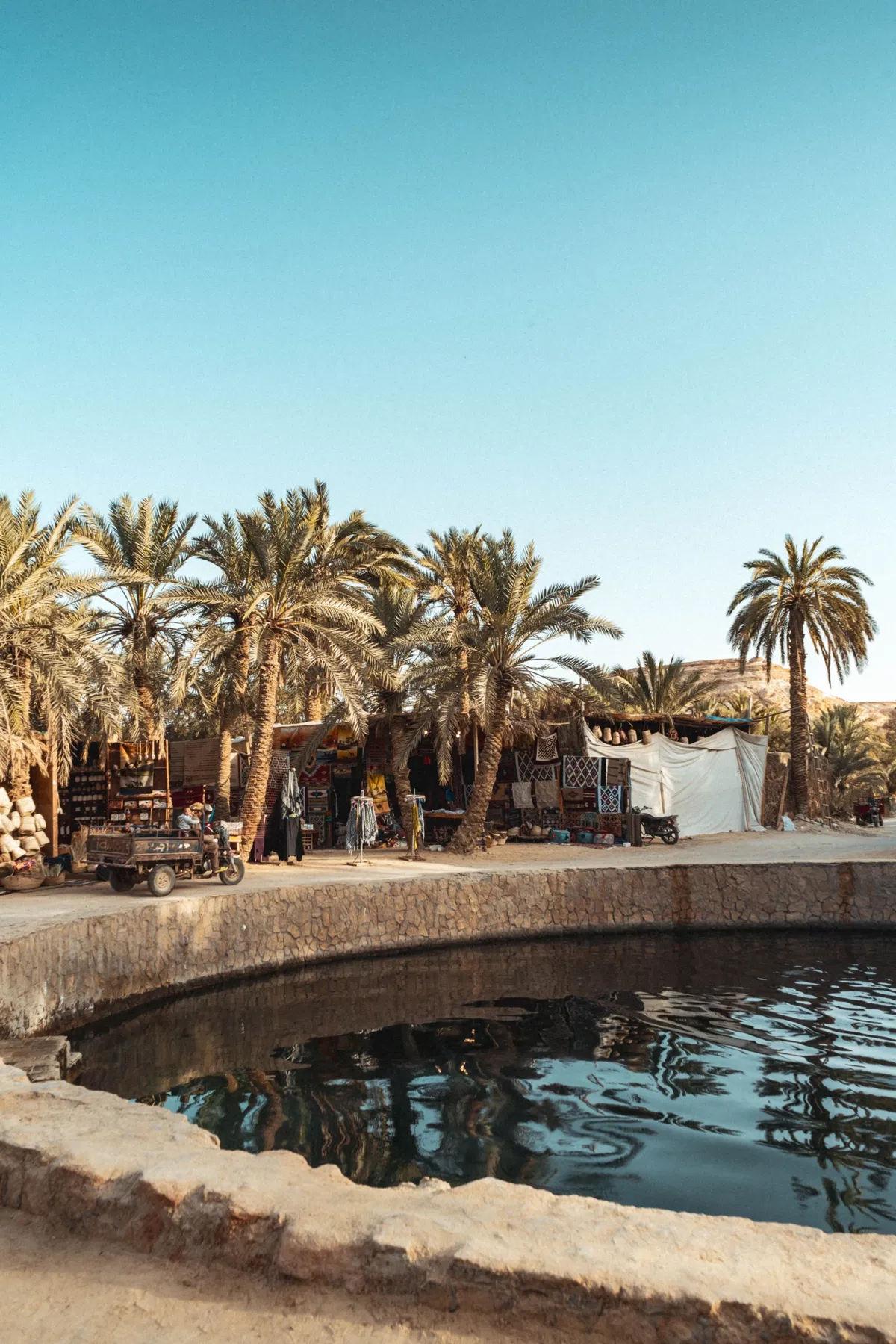 A round stone-edged spring pool in Siwa, with palm trees and market stalls selling crafts behind it.