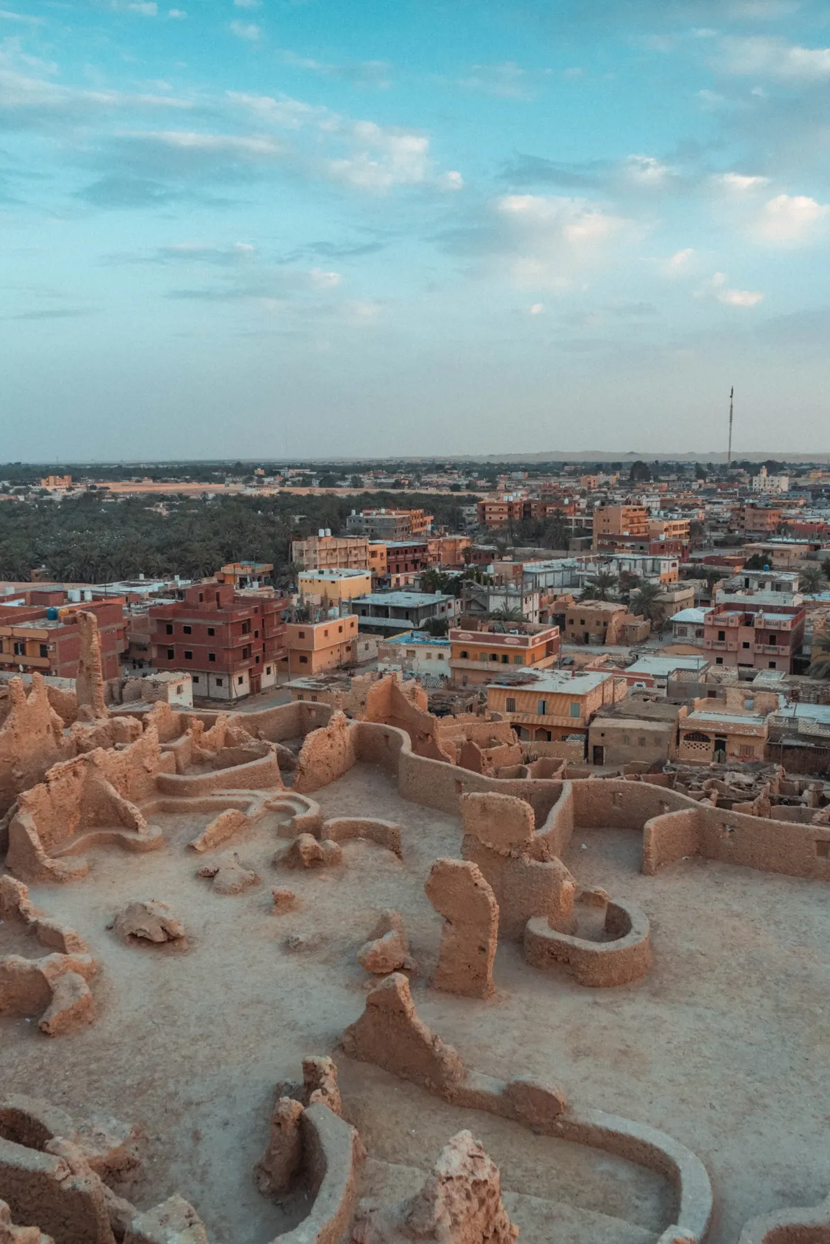 Crumbling mud-brick ruins in the foreground above Siwa's brick and concrete houses and palm groves in soft evening light.