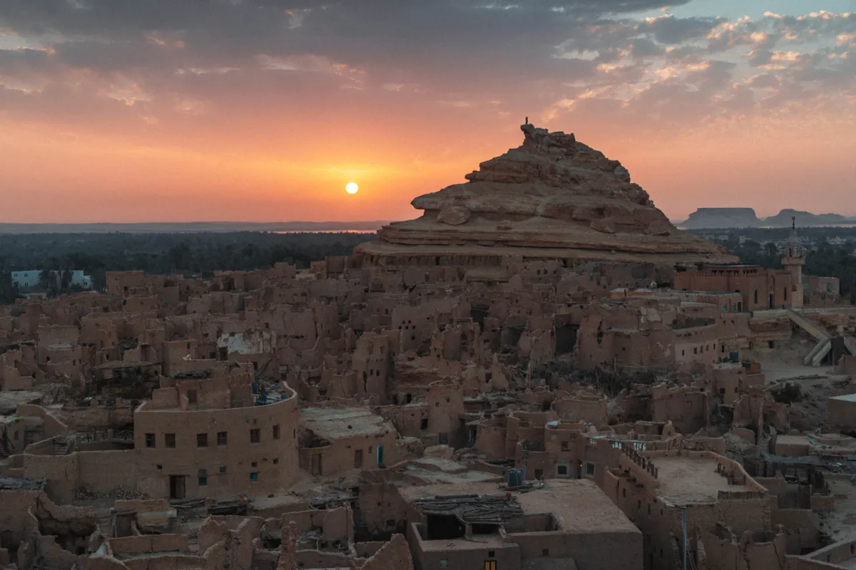 The sun sets in an orange sky behind the mud-brick houses of Siwa's old town, which climb a rocky hill.