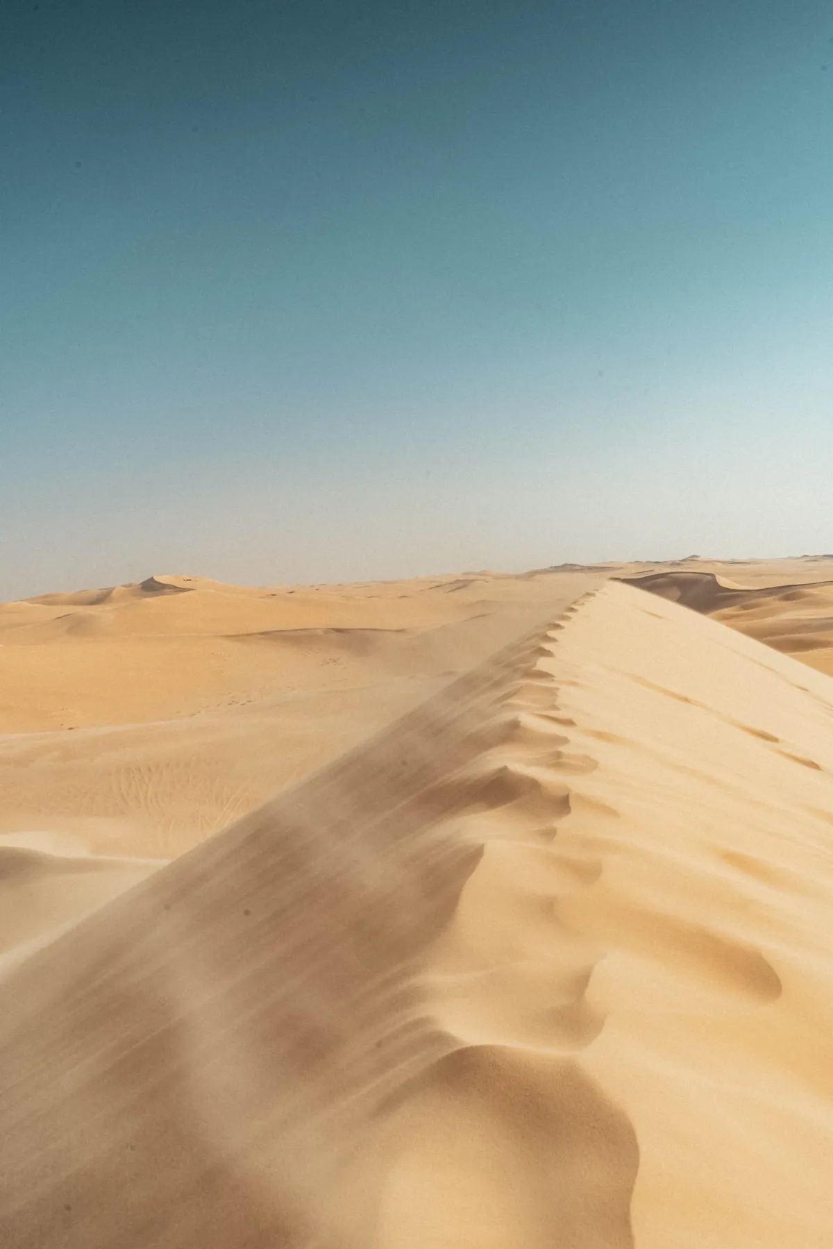 A sharp wind-blown sand dune crest leads into endless dunes under a hazy teal sky near Siwa.