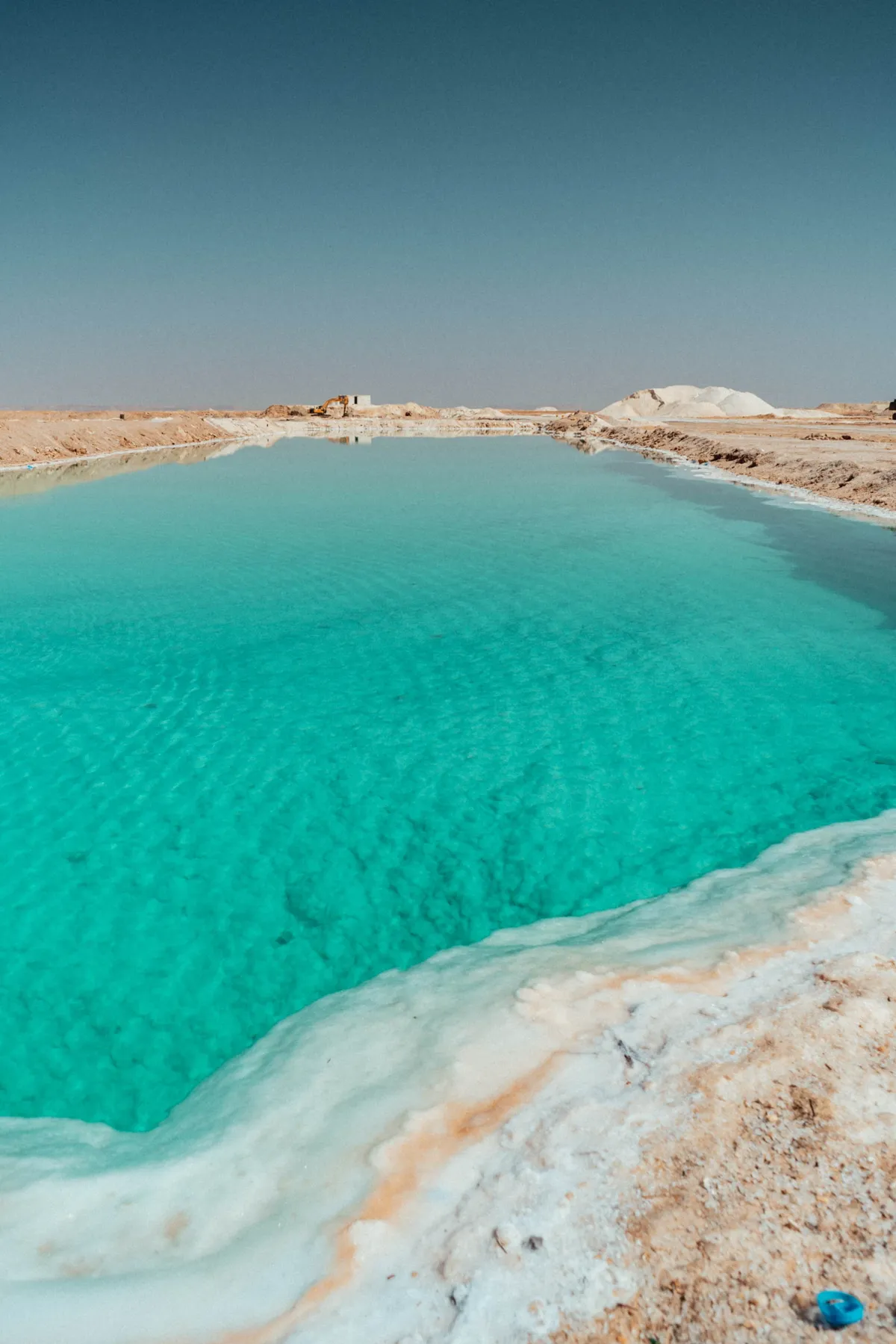 A turquoise salt pool edged with white salt crust lies in the desert near Siwa, with a salt mound beyond.