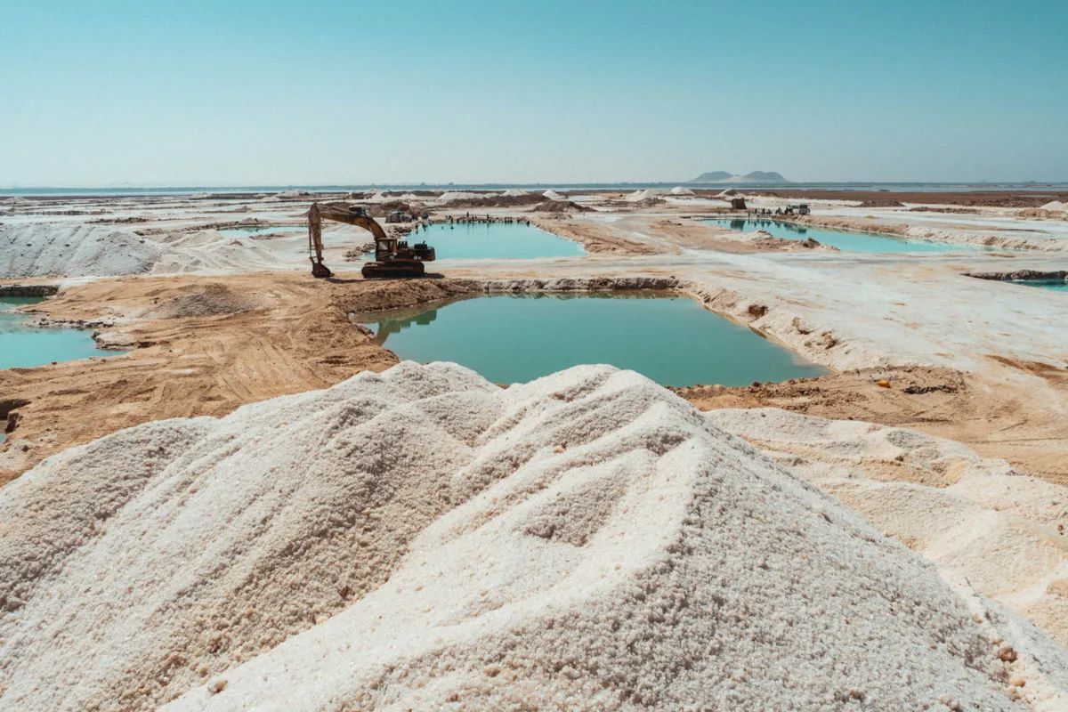 A large pile of white salt in the foreground, with an excavator and green-blue brine pools in the flats beyond.