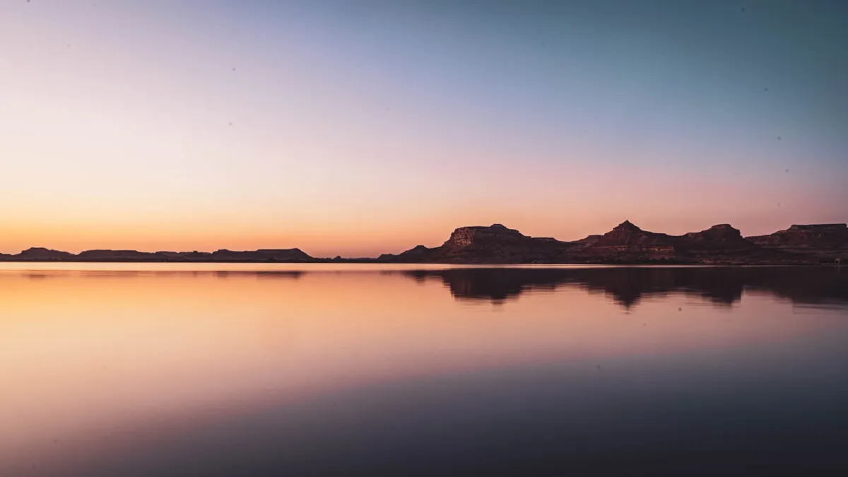 A calm lake at dusk reflects the pink and orange sky and silhouetted rock hills near Siwa.