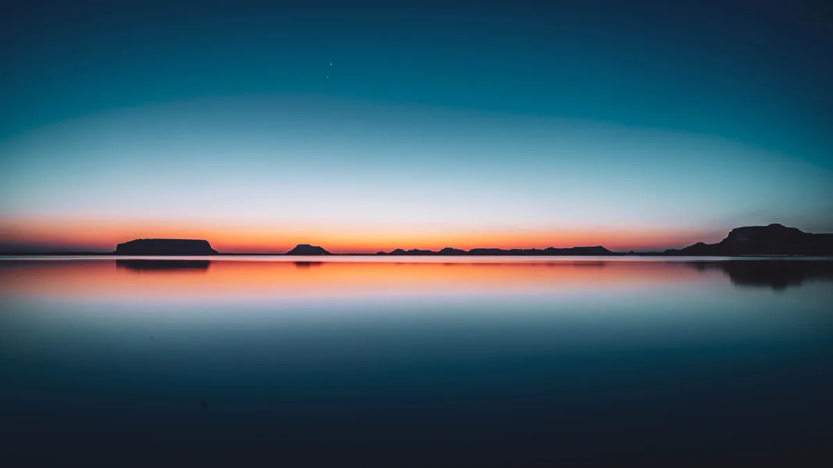 A mirror-still lake at blue hour, with a thin orange glow on the horizon behind dark flat-topped hills.