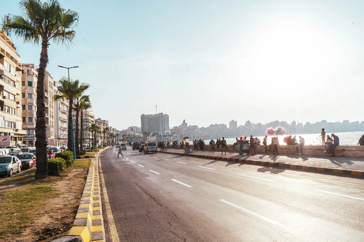 The Alexandria Corniche runs along the sea in late light, with palm trees, apartment blocks and people on the seawall.