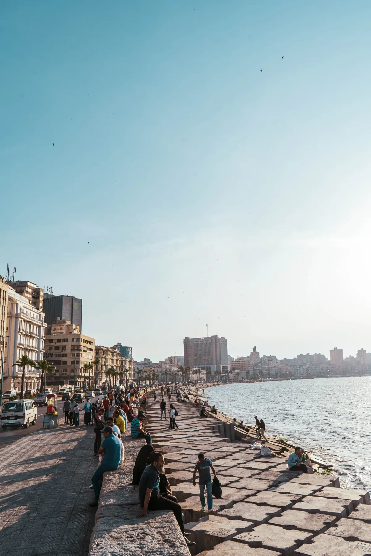 People sit along the stone breakwater of the Alexandria Corniche, with the curving coastline and city buildings fading into haze.
