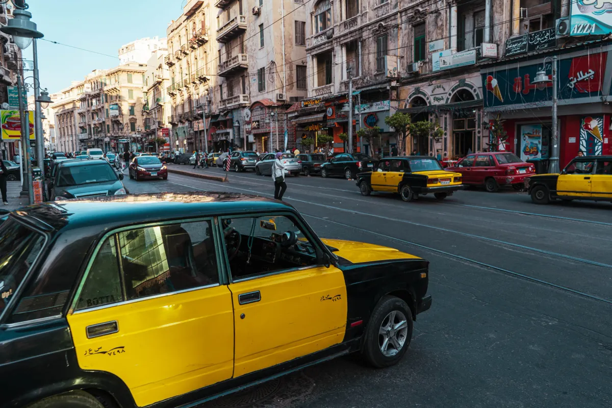 A yellow and black taxi in the foreground on a busy Alexandria street lined with old apartment buildings and shops.