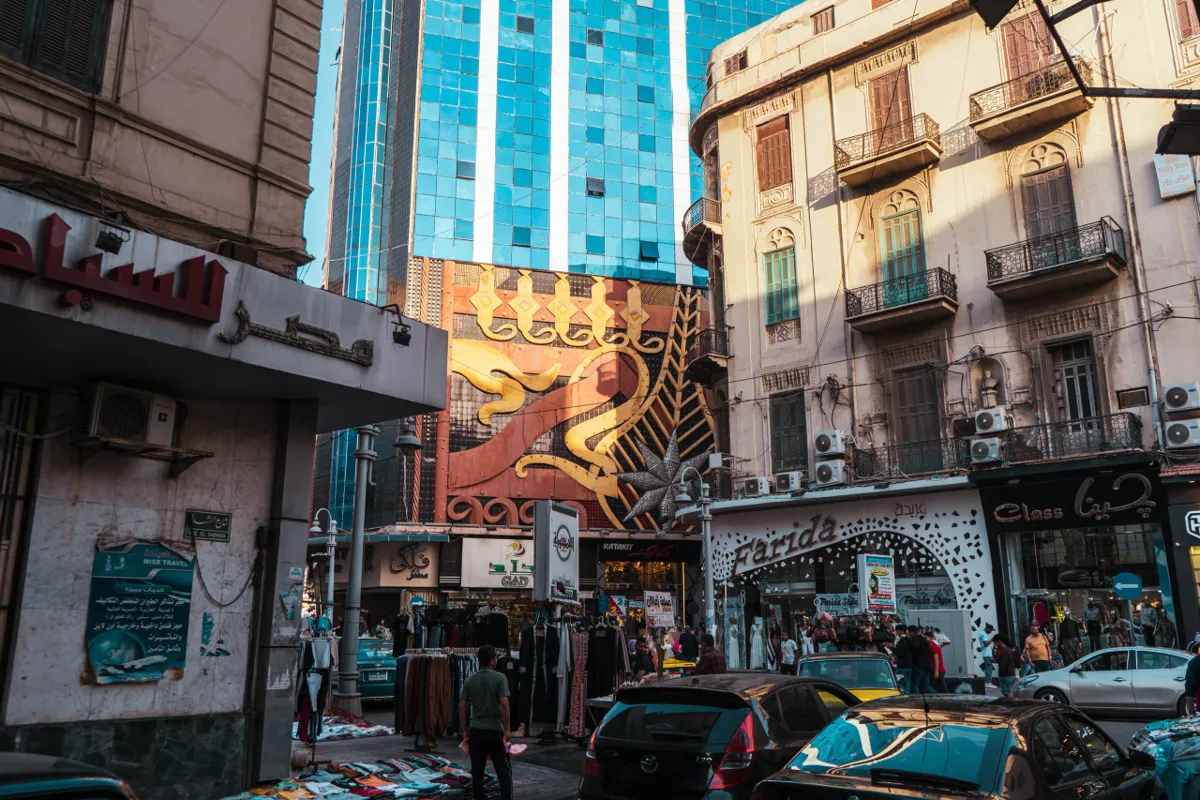 An Alexandria street corner with shops, vendors and a decorated facade, with a glass tower rising behind older balconied buildings.