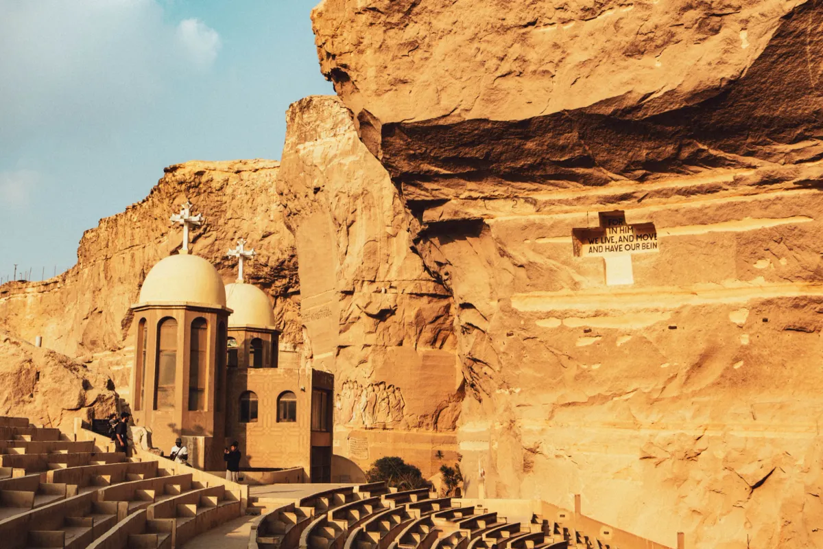 A sandstone cliff with a rock-cut church, small domes with crosses and curved stone seating in warm light in Cairo.