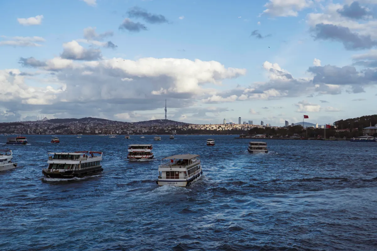 Several ferries cross the choppy blue Bosphorus under a cloudy sky, with a hillside skyline and TV tower beyond.