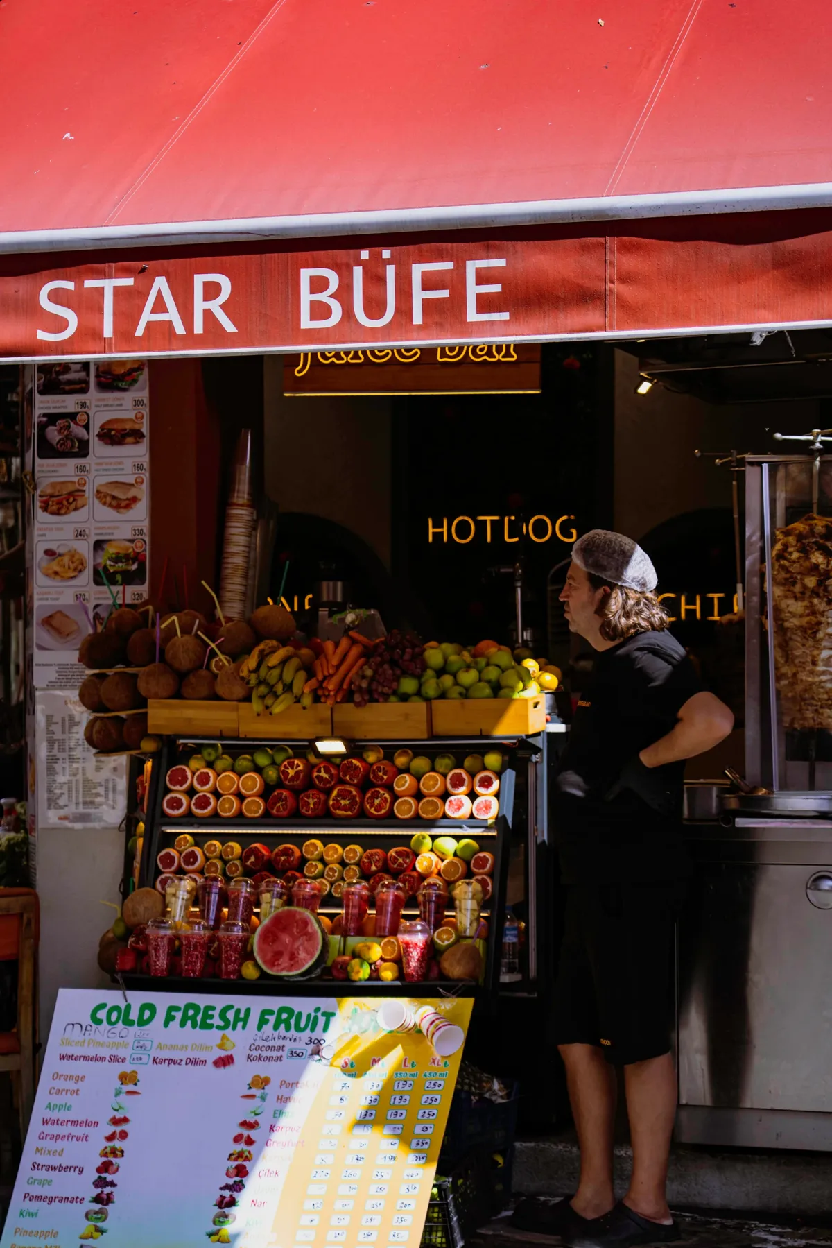 A man in a cap stands at a fresh juice and street food stall with a red awning in Istanbul.