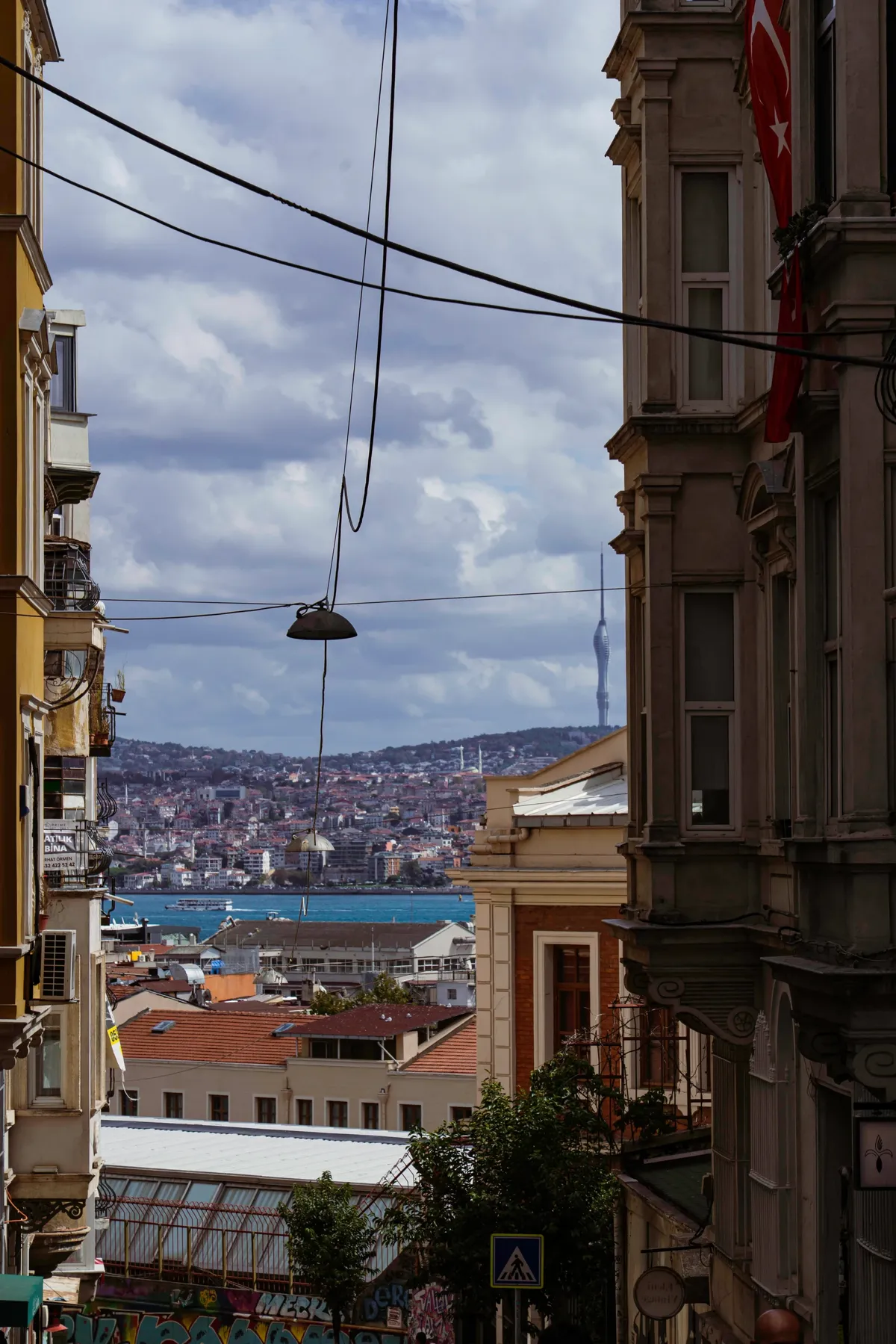 A narrow Istanbul street between old buildings opens onto the Bosphorus, a hillside district and a tall TV tower.