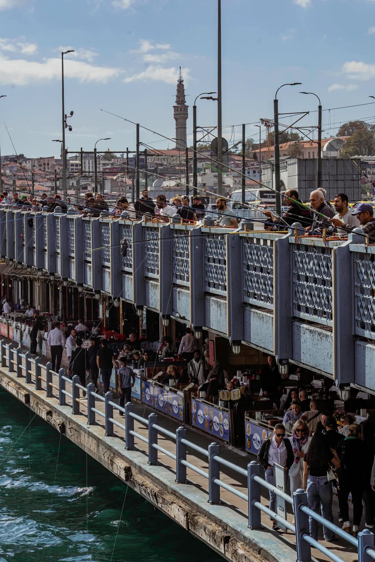 Crowds of people fish along the railing of the Galata Bridge in Istanbul, with restaurants below and the city behind.