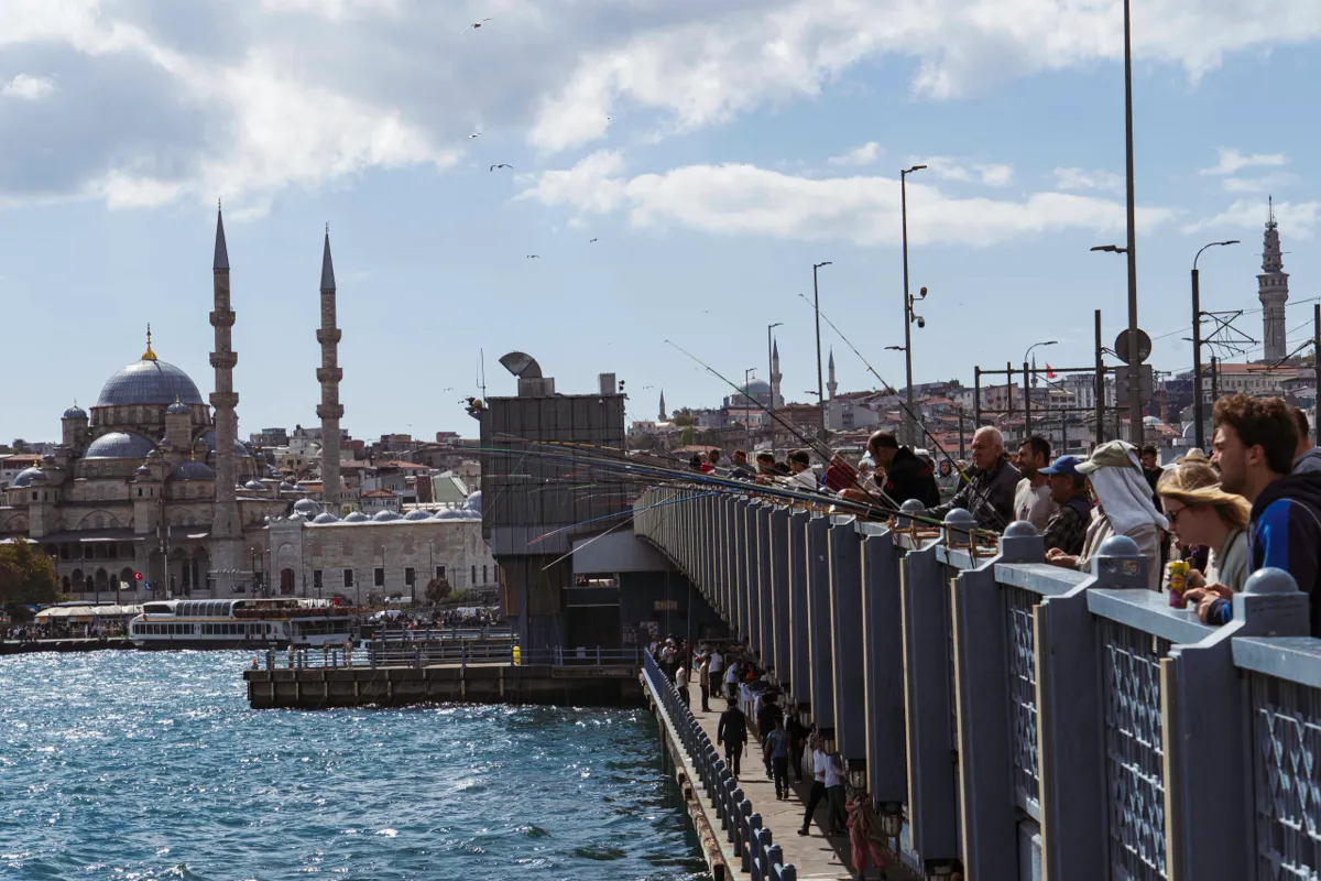 Fishermen line the Galata Bridge railing in Istanbul, with the Bosphorus at left and a domed mosque across the water.