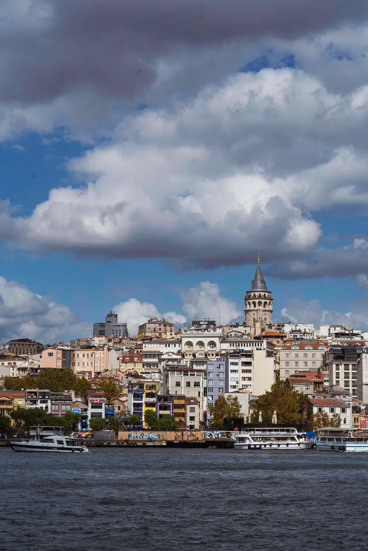 The Galata Tower rises above hillside buildings across the water in Istanbul, with ferries below and big clouds above.