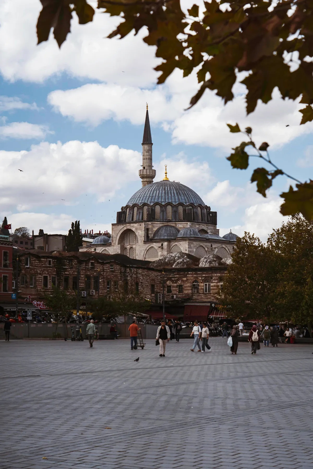 A domed mosque with one minaret stands beyond a paved plaza where people walk, framed by autumn leaves in Istanbul.
