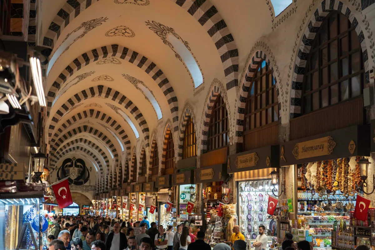 A crowded vaulted bazaar arcade in Istanbul with striped stone arches, shop signs and red Turkish flags.