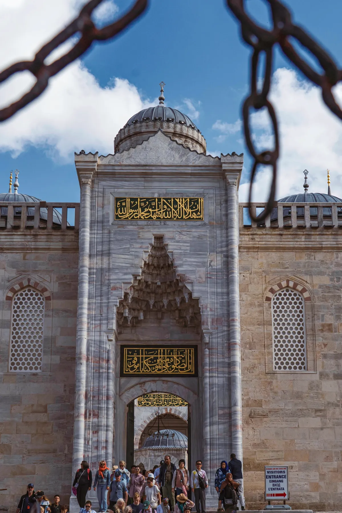 Visitors gather at the marble gateway of a mosque in Istanbul, with gold calligraphy panels and domes above.
