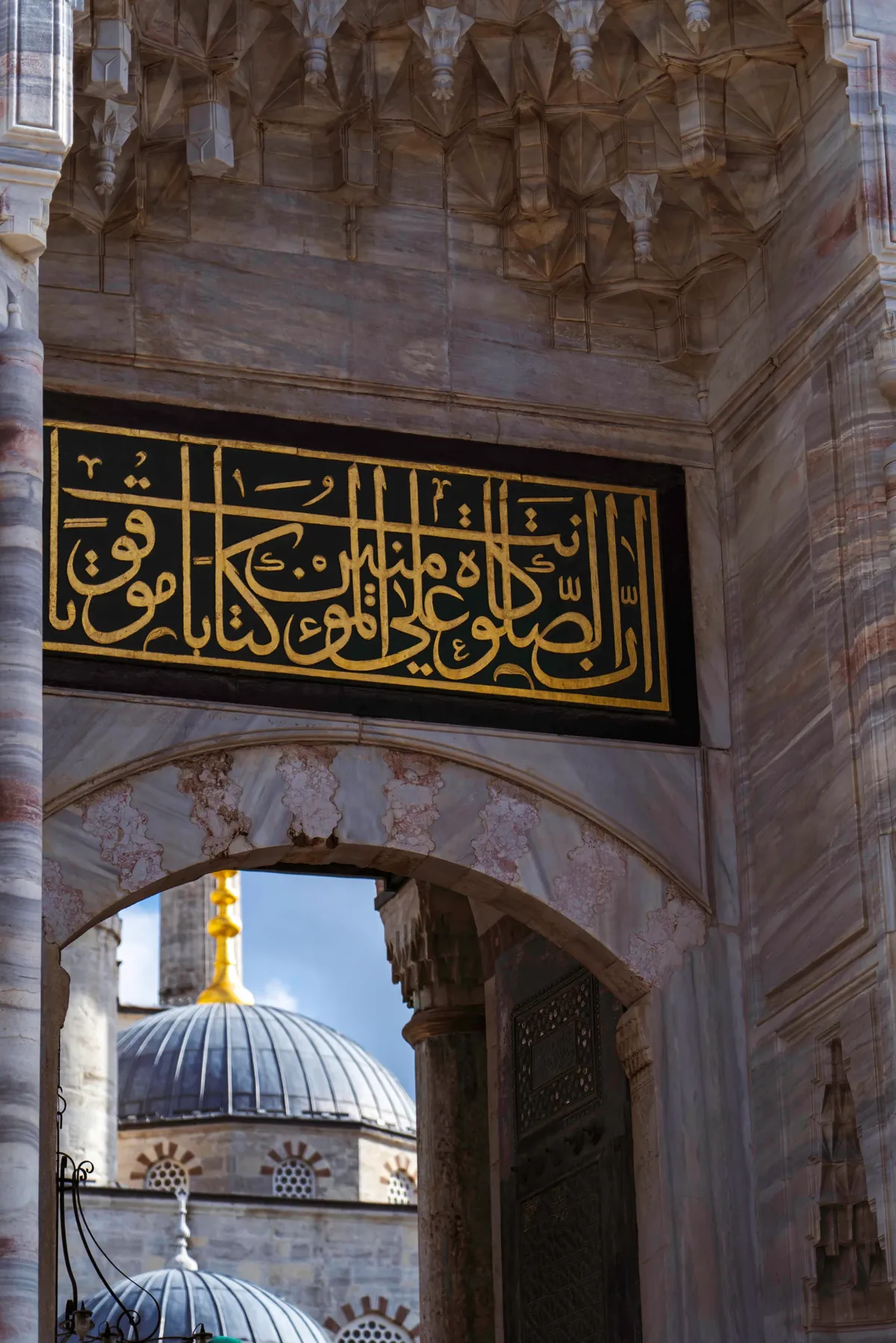 A black and gold calligraphy panel and carved stone arch at a mosque gate, with domes beyond in Istanbul.