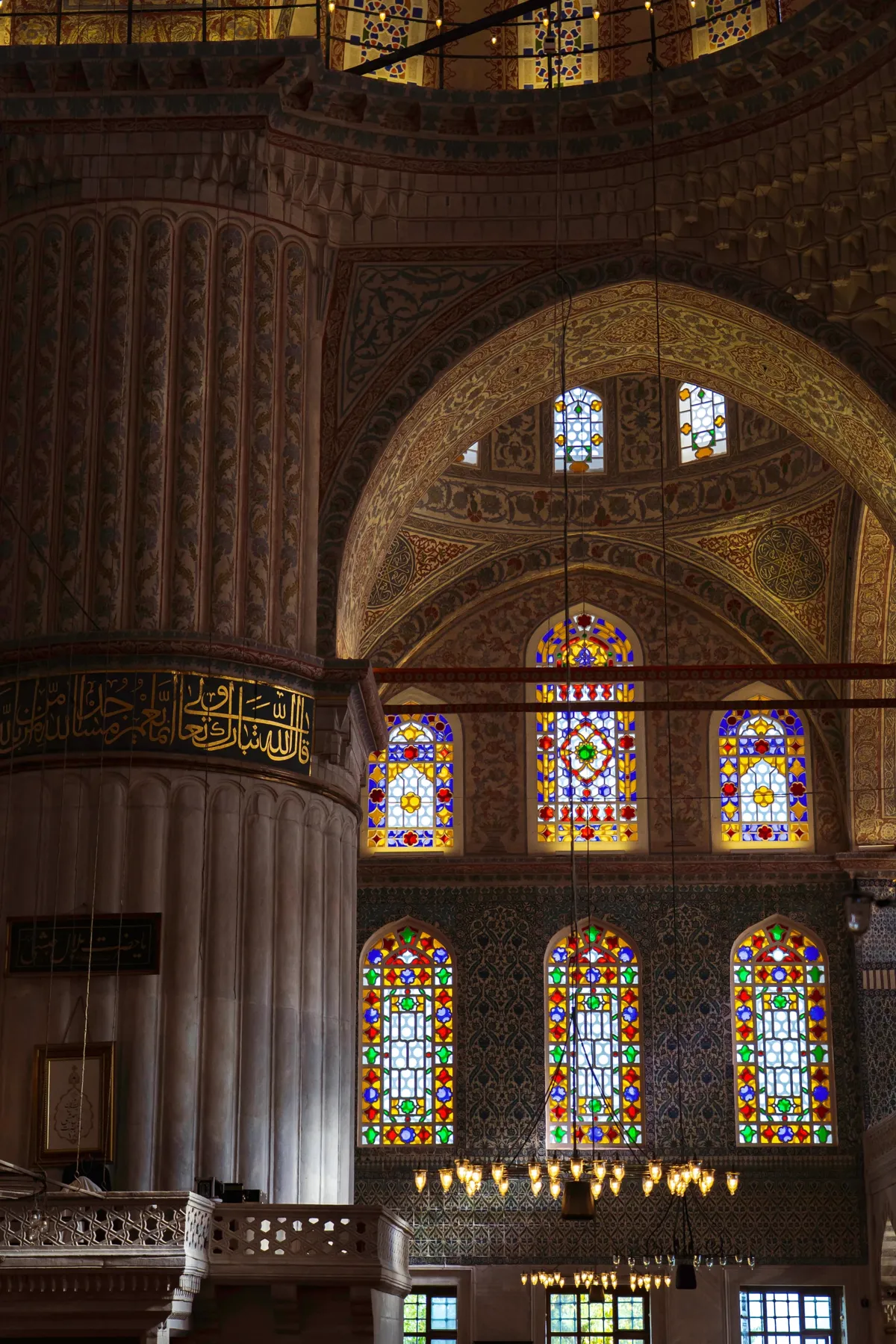 Tall stained glass windows and painted tilework inside a mosque in Istanbul, with hanging lamps below.