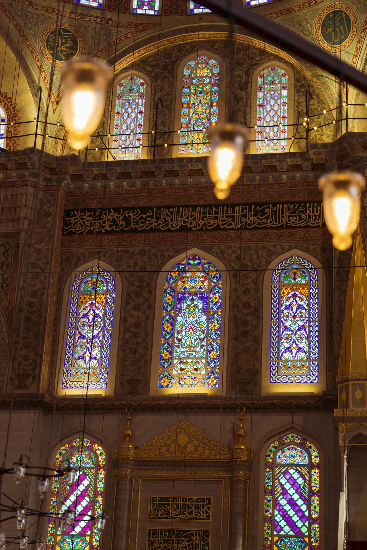 Stained glass windows, gold calligraphy and glowing hanging lamps inside a mosque in Istanbul.