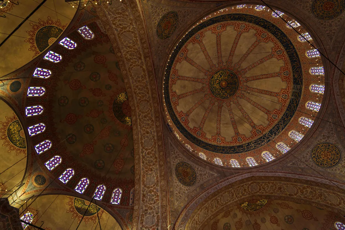 Looking up at a mosque's painted central dome, with a ring of small windows and ornate arabesque patterns.