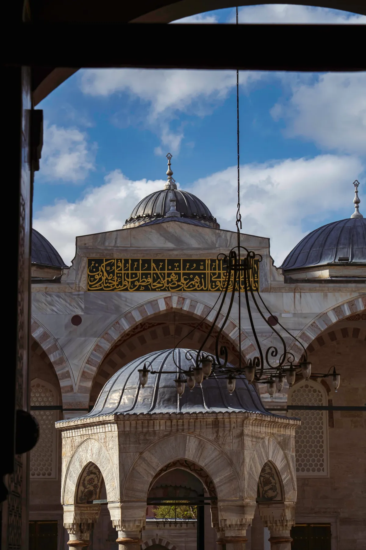 A hanging lamp frames a mosque courtyard with a domed ablution fountain and calligraphy above, seen from a dark doorway.